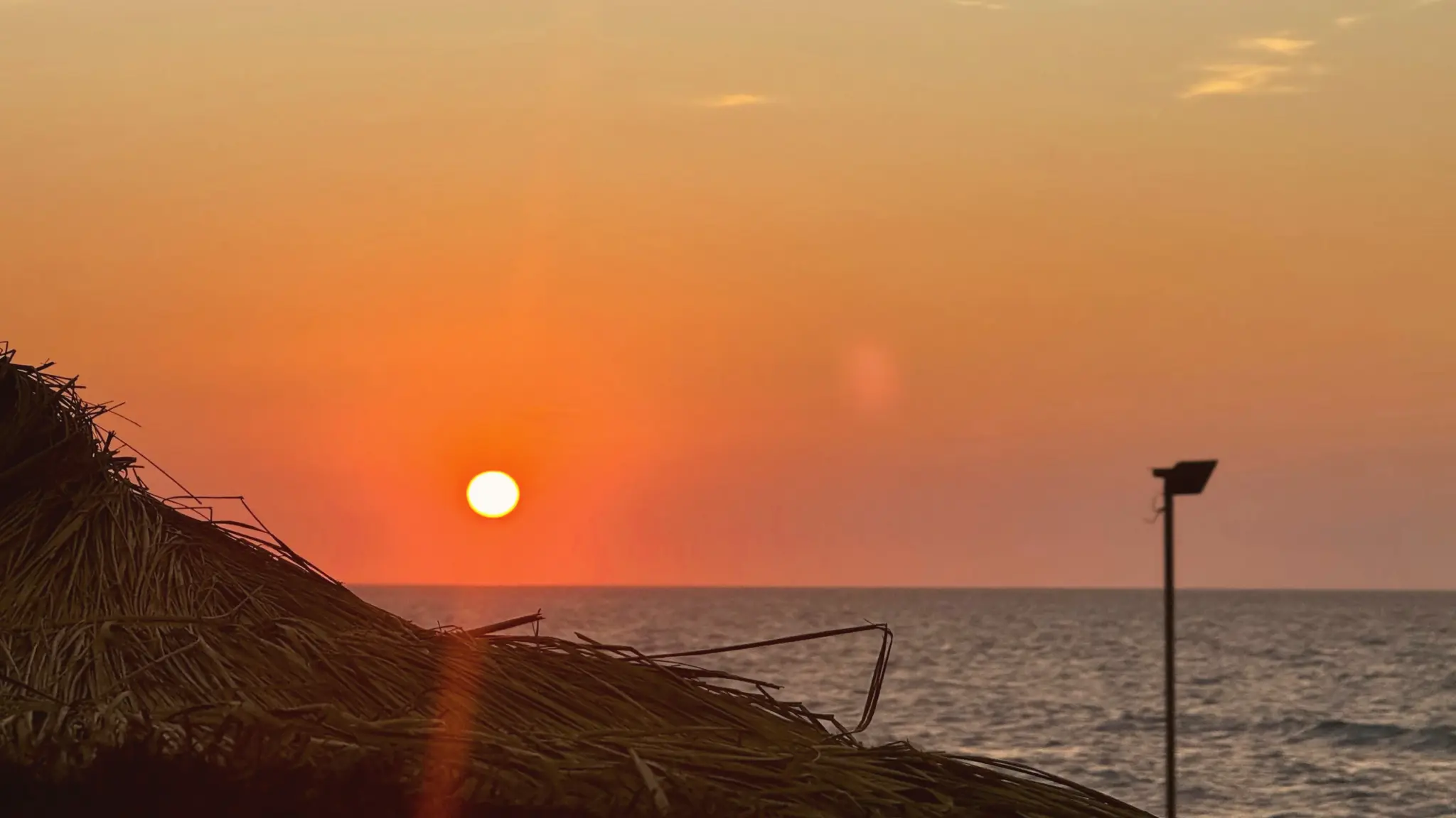 Orange sun setting over the Mediterranean beyond a thatched parasol and lamp post