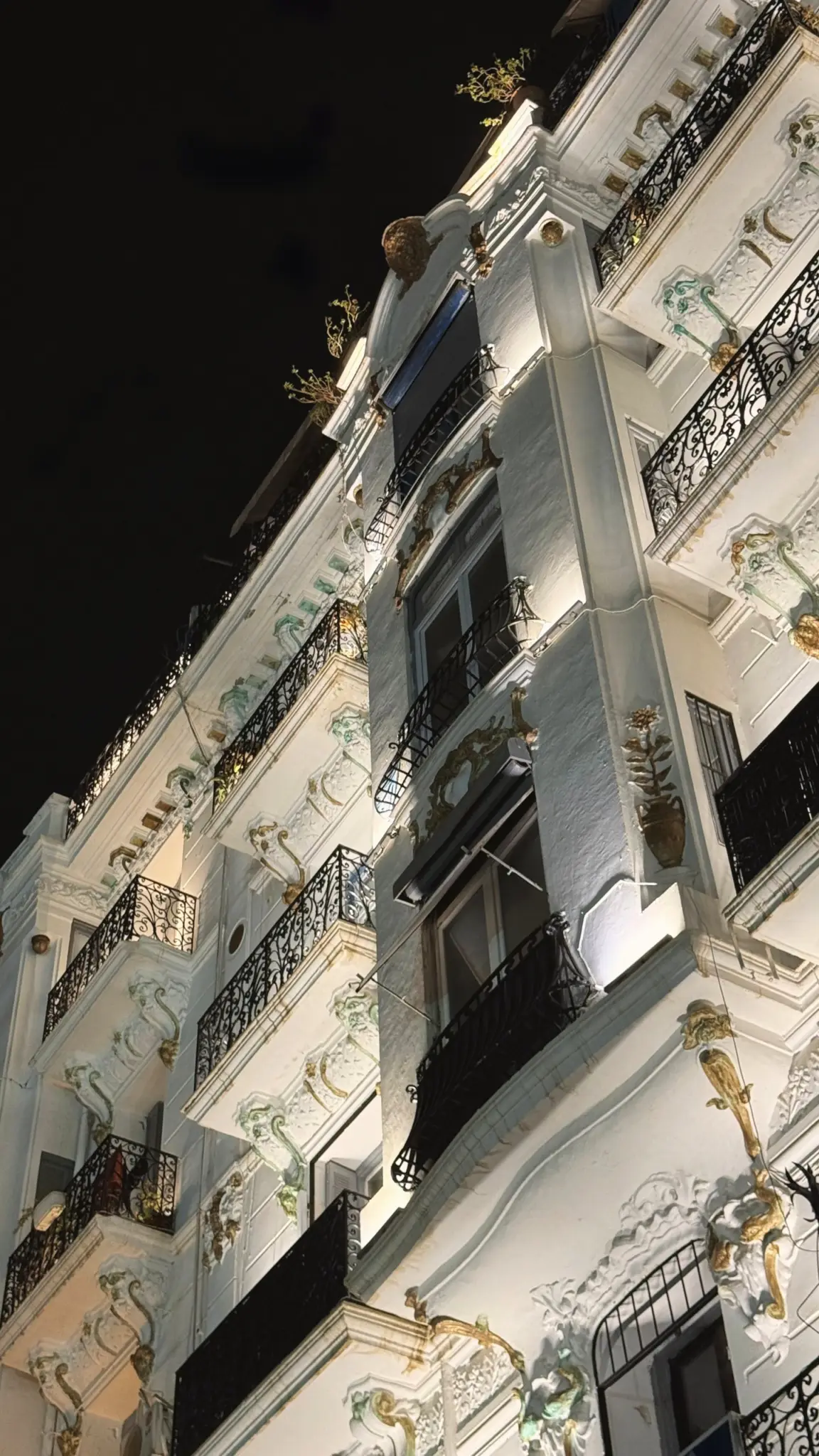 Looking up at an illuminated white facade with wrought-iron balconies at night