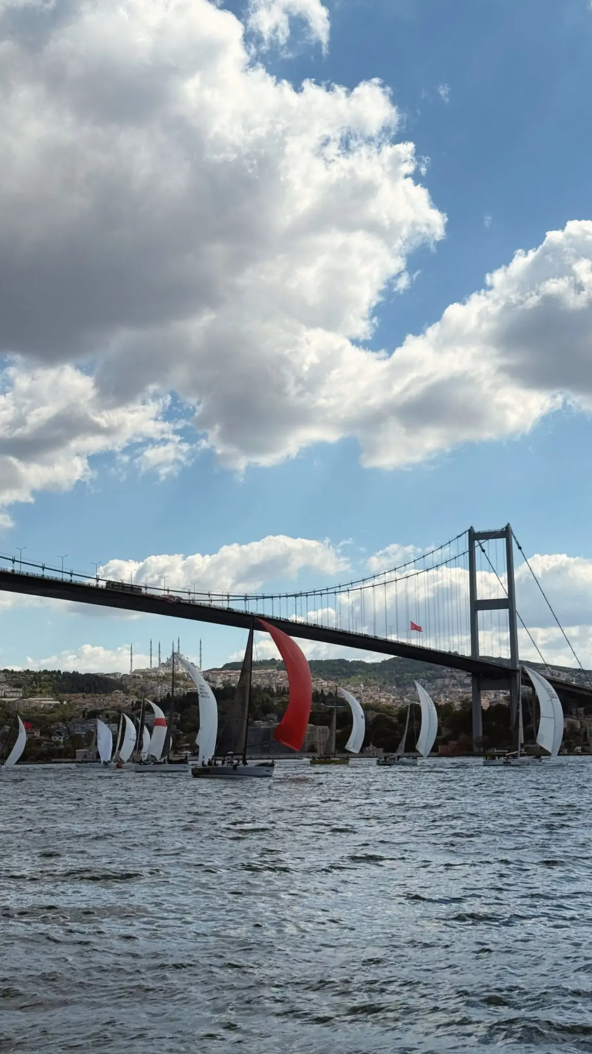 Sailing yachts racing beneath the Bosphorus Bridge under billowing clouds