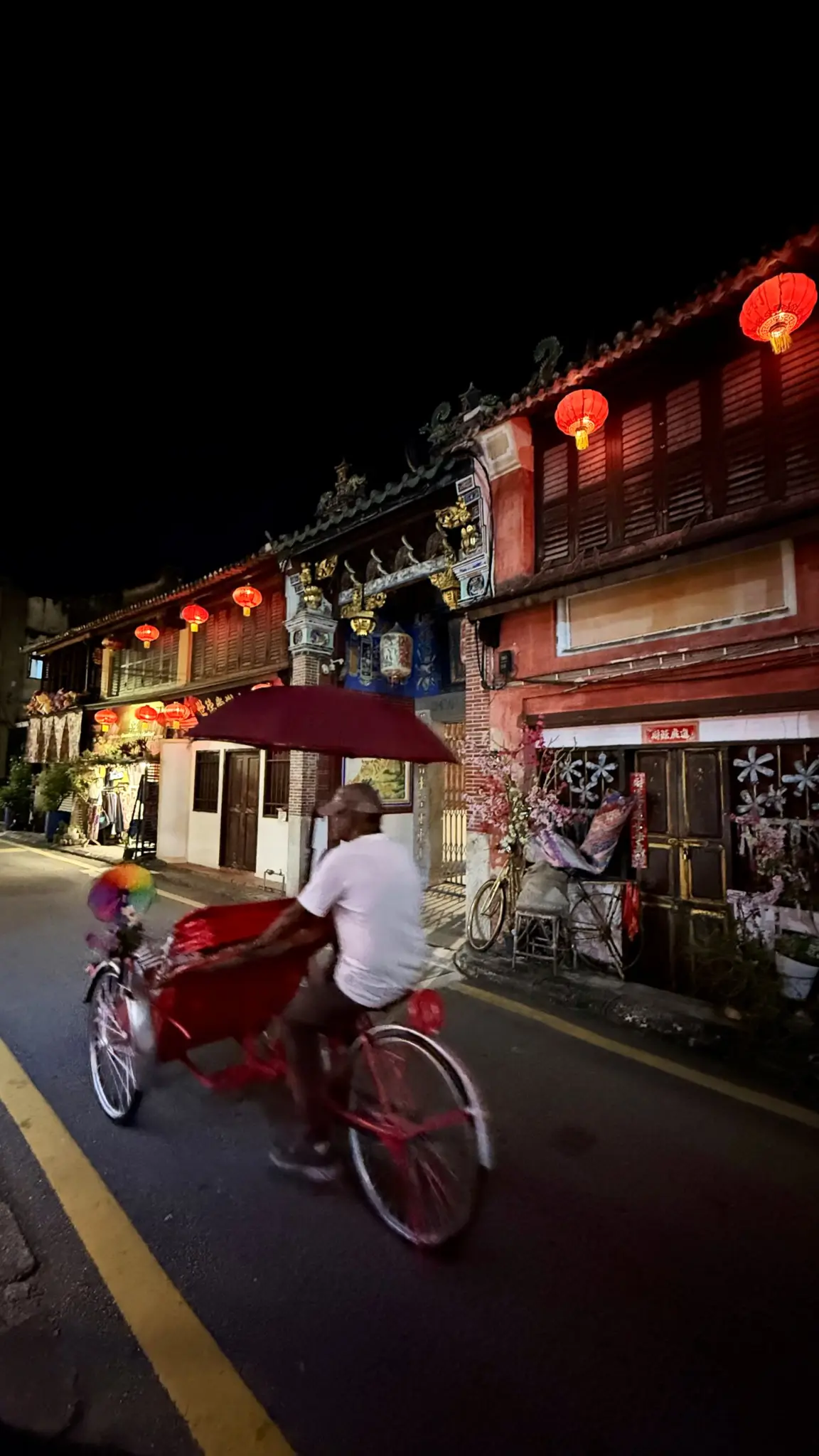 Trishaw rider pedalling past lantern-lit shophouses at night